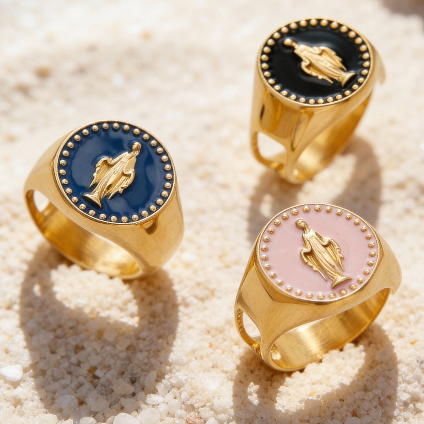 Three gold spiritual rings with raised figure and enamel tops in navy, black, and pink on sand
