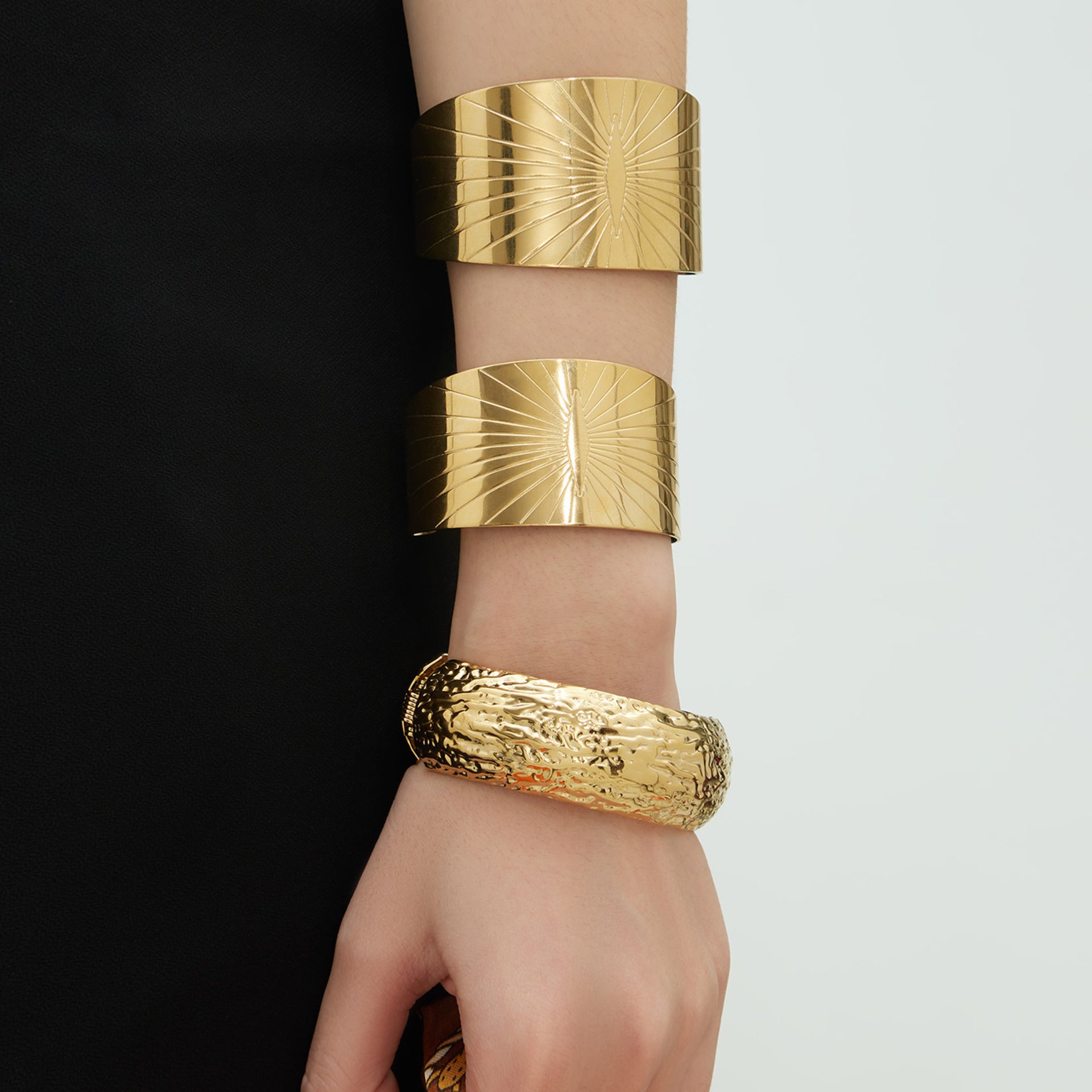 Woman's arm wearing two wide gold cuffs with sunburst design and a hammered gold bangle on white background